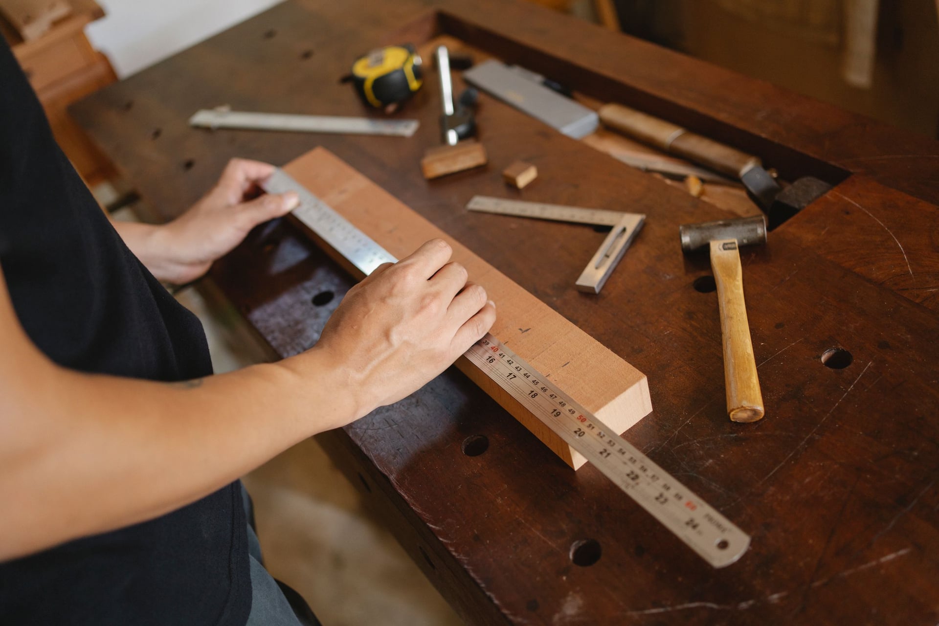 Hands measuring a wooden board with a steel ruler on a workbench.