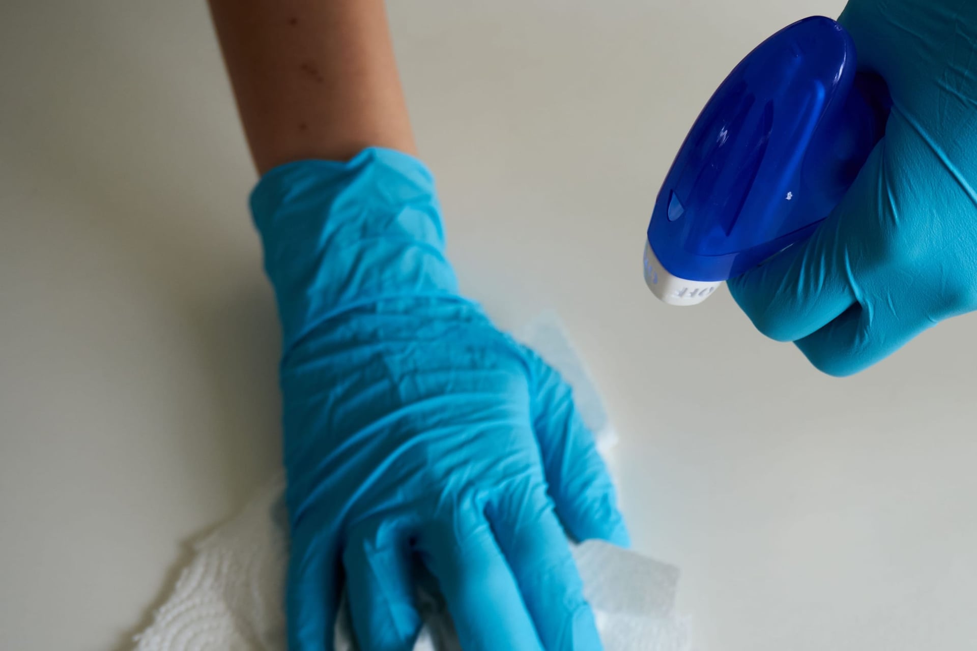 Gloved hands wiping a white desk with disinfectant spray.